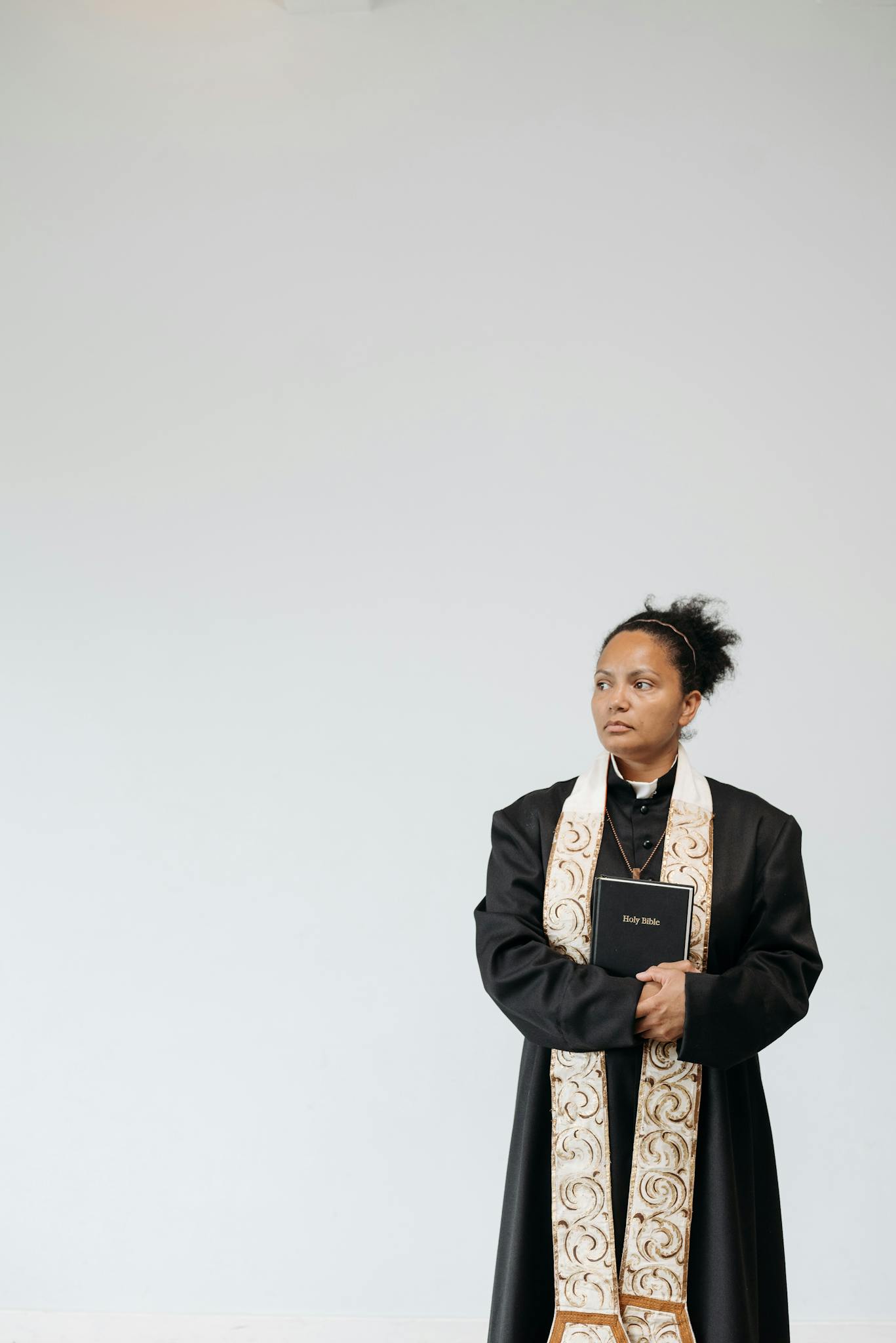 A female pastor standing indoors holding a Bible, wearing a cassock and stole.