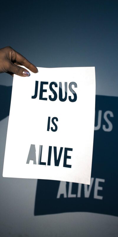 A person holding a white paper with the message 'Jesus is Alive' against a blue background.